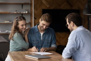 Happy millennial couple signing property purchase contract, insurance agreement, filling bank application for mortgage, buying house, loan. Young married man and woman meeting with legal expert