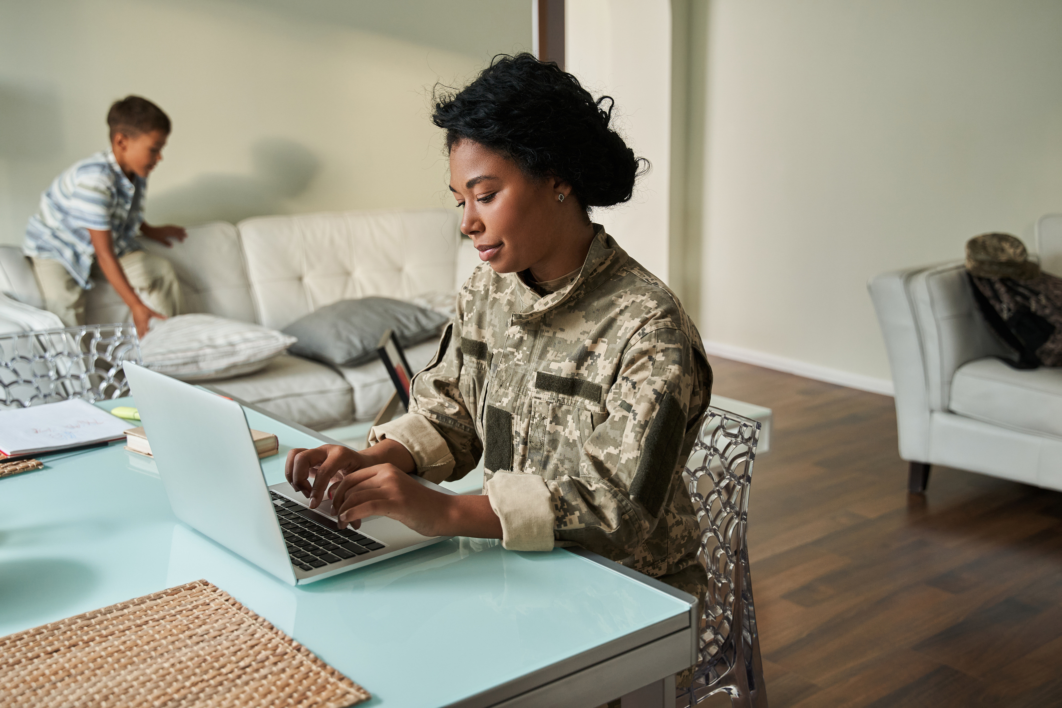 Serious black woman in camouflage uniform using laptop while sitting at table. After war rehabilitation of female soldier. Little boy play on couch in background. Modern comfortable flat